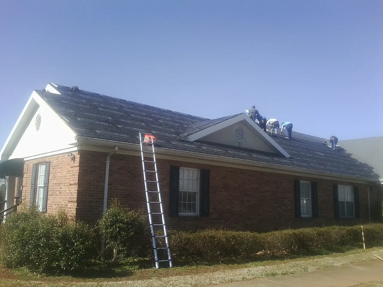Crew tearing shingles off a brick ranch roof with a ladder set — Metal Roof Repair work by our crew in Pelahatchie