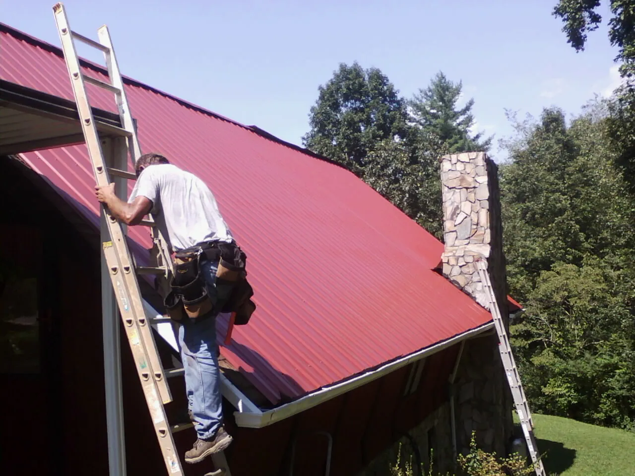 Roofer on a ladder beside a red metal roof and stone chimney — Soffit & Fascia Repair by Roofing Brothers in Pelahatchie